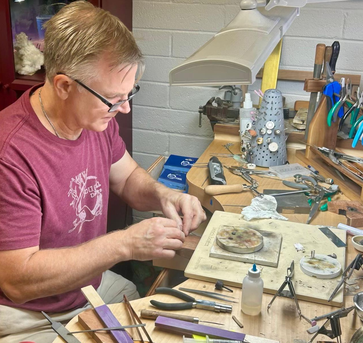 A maker at the jeweller's bench, hands over a piece under the lamp, pliers and files laid out around him.