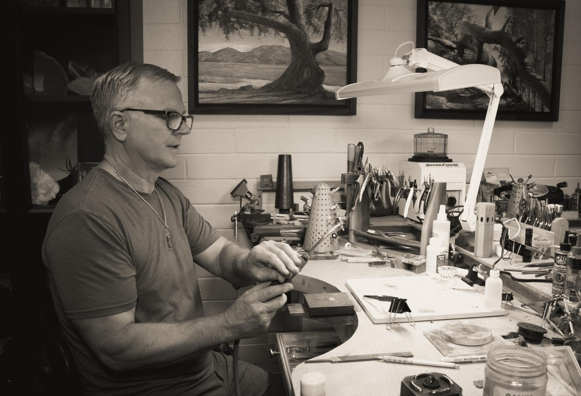 Jeremy at the jeweler's bench, working a piece under the lamp, with two of Les's paintings on the wall behind him.