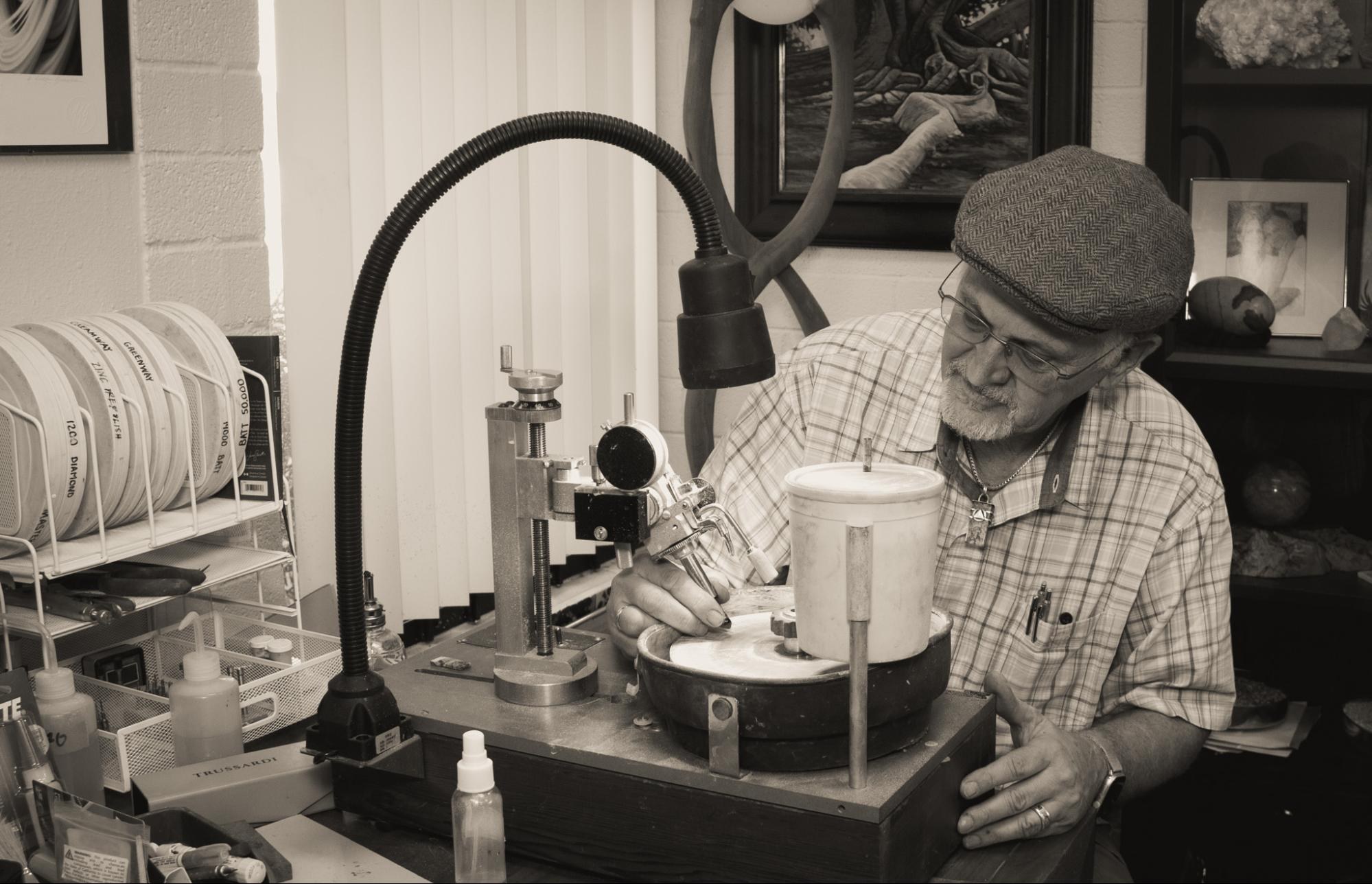 Les in his flat cap at the faceting machine, working a stone under the lamp, one of his paintings on the wall behind him.