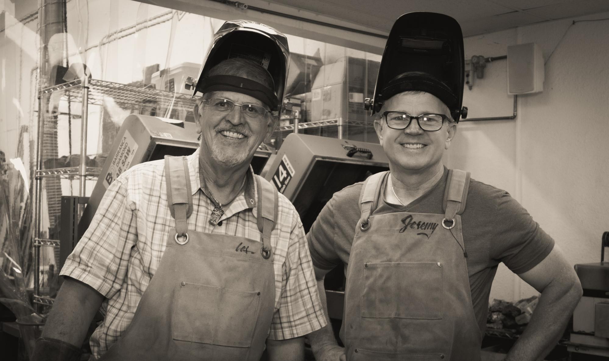 Les and Jeremy side by side in the workshop, welding hoods pushed up and aprons on, smiling, with the slab saws behind them.
