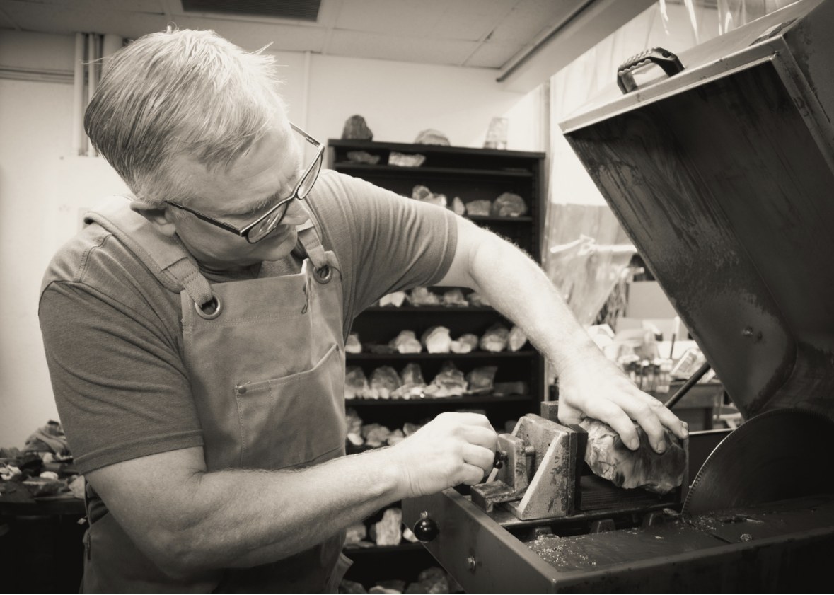 Jeremy at the slab saw in his apron, setting a rough stone in the vise before the first cut, shelves of rough behind him.